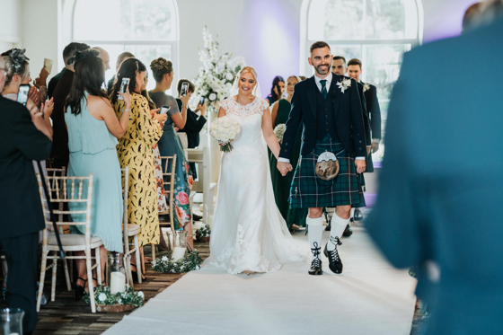 Bride and groom smile as they walk hand in hand back down the aisle at The Venue, Eskmills
